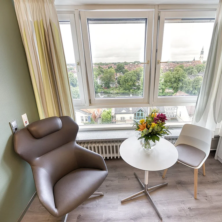 Zwei Stühle stehen bei einem Tisch vor einem großen Fenster. Auf dem Tisch steht ein Blumenstrauß in einer Vase. Rechts im Bild steht ein weißer Schrank.