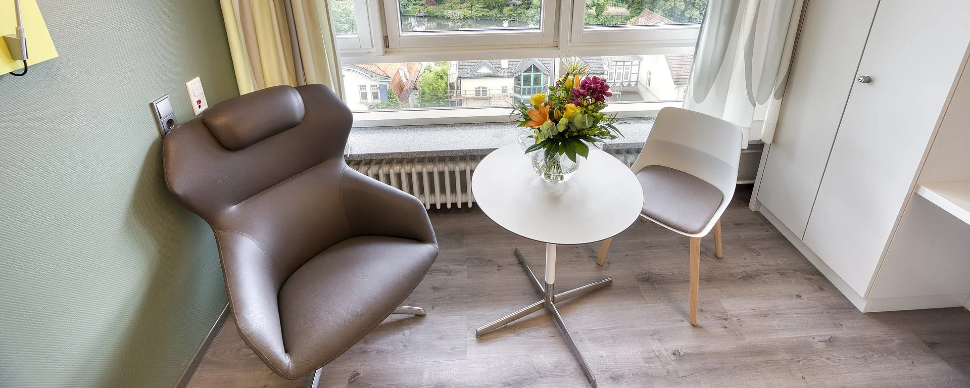 Zwei Stühle stehen bei einem Tisch vor einem großen Fenster. Auf dem Tisch steht ein Blumenstrauß in einer Vase. Rechts im Bild steht ein weißer Schrank.