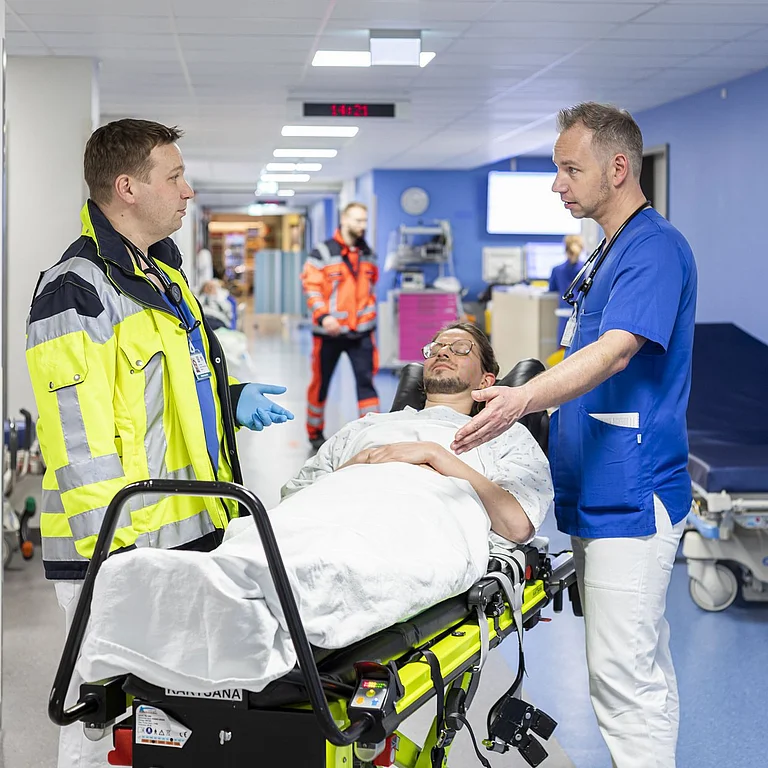 Ein Notarzt unterhält sich mit einem Pfleger. Zwischen den beiden liegt eine Patientin auf einer Trage.