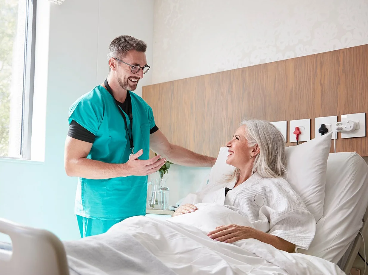 Ein Pfleger spricht freundlich mit einer älteren Patientin, die in einem modernen Krankenhausbett liegt. Das helle Zimmer wirkt ruhig und einladend.