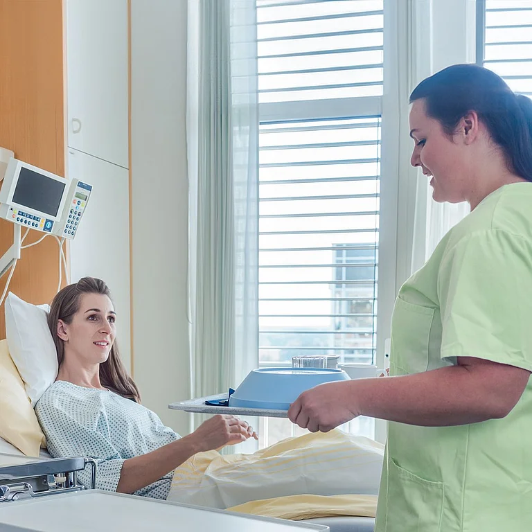 Eine Pflegekraft überreicht einer Patientin im Krankenhausbett Unterlagen – freundlicher Austausch auf der Station.