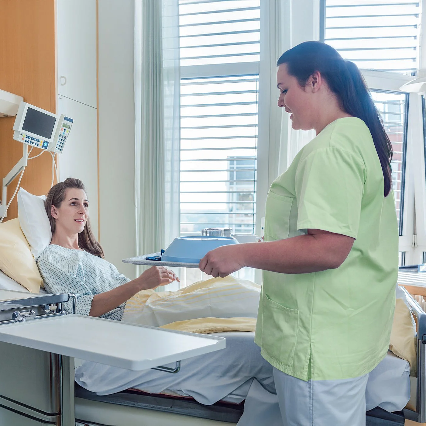 Eine Pflegekraft überreicht einer Patientin im Krankenhausbett Unterlagen – freundlicher Austausch auf der Station.