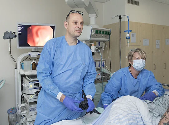 Ärzteteam in Schutzkleidung führt endoskopische Untersuchung bei Patient im Behandlungszimmer mit Monitor durch.