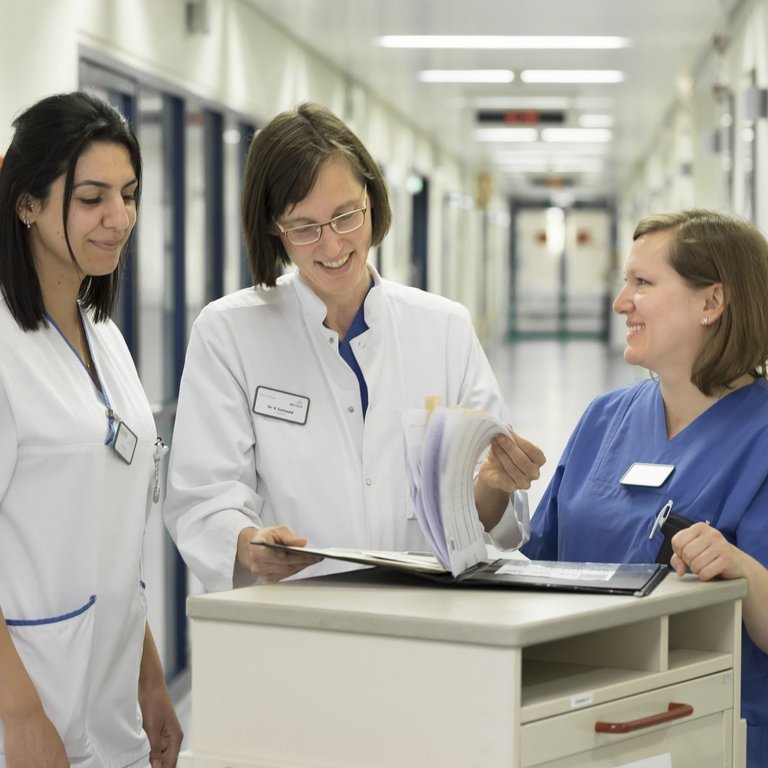 Zwei Ärztinnen und eine Pflegerin stehen an einem Rollcontainer, auf dem eine Patientenmappe steht und unterhalten sich.