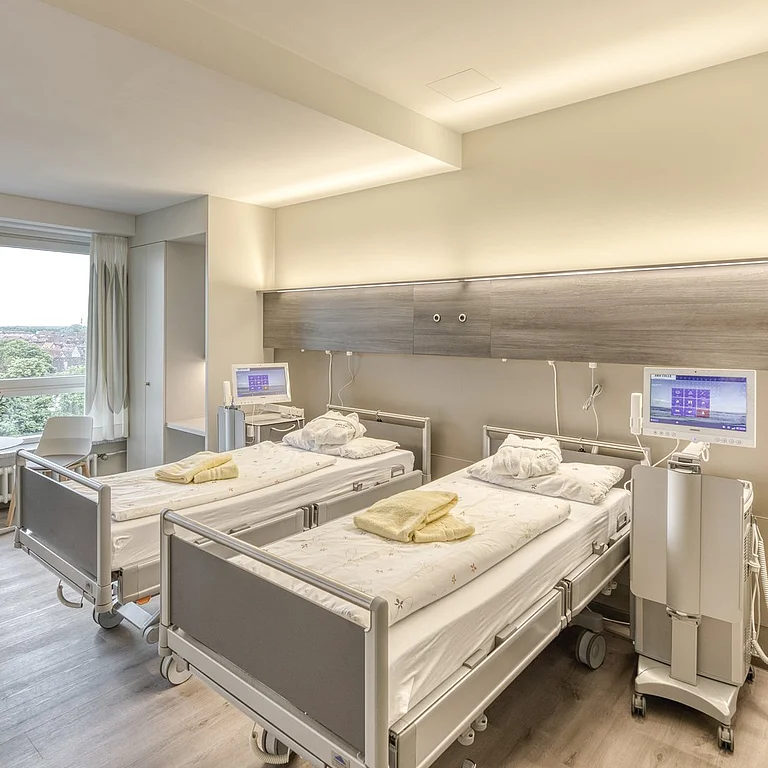 Das Bild zeigt ein modernes, hell eingerichtetes Patientenzimmer mit zwei Krankenhausbetten, medizinischen Geräten und großem Fenster mit Ausblick ins Grüne.