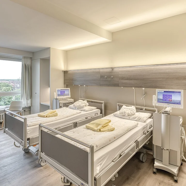 Das Bild zeigt ein modernes, hell eingerichtetes Patientenzimmer mit zwei Krankenhausbetten, medizinischen Geräten und großem Fenster mit Ausblick ins Grüne.