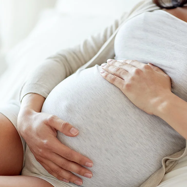 Schwangere Frau hält schützend ihre Hände auf dem Babybauch im Sitzen