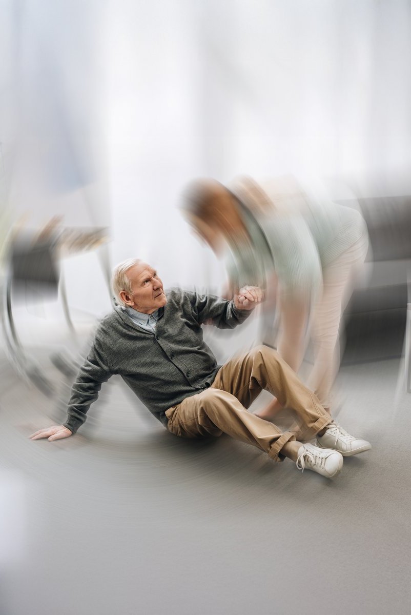 Eine Pflegekraft hilft einem älteren Mann, der in einem Aufenthaltsraum gestürzt ist, wieder auf die Beine. Die Szene ist leicht verschwommen, vermittelt aber Fürsorge und schnelle Hilfe.