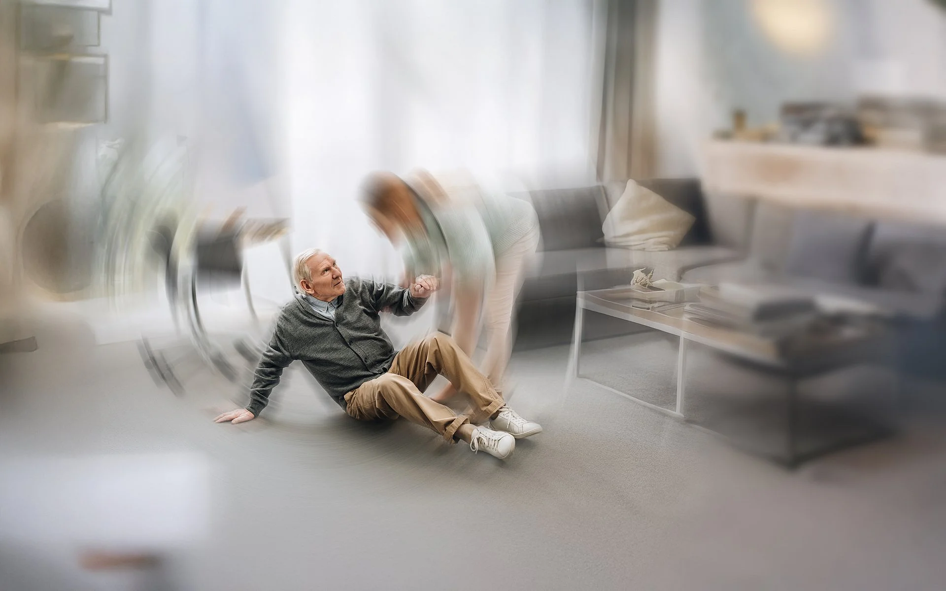 Eine Pflegekraft hilft einem älteren Mann, der in einem Aufenthaltsraum gestürzt ist, wieder auf die Beine. Die Szene ist leicht verschwommen, vermittelt aber Fürsorge und schnelle Hilfe.