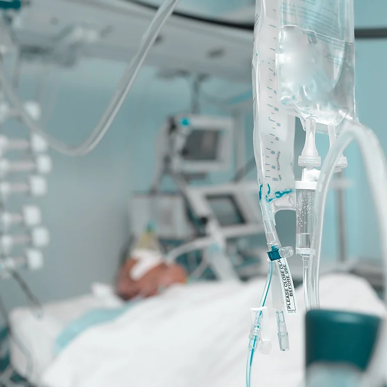 Nahaufnahme einer Infusion im Vordergrund, im Hintergrund verschwommen ein Patient auf der Intensivstation.