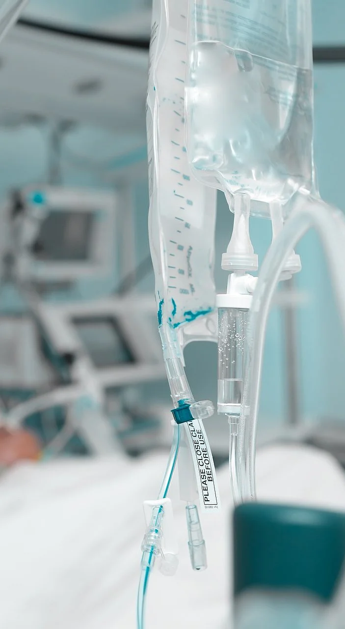 Nahaufnahme einer Infusion im Vordergrund, im Hintergrund verschwommen ein Patient auf der Intensivstation.