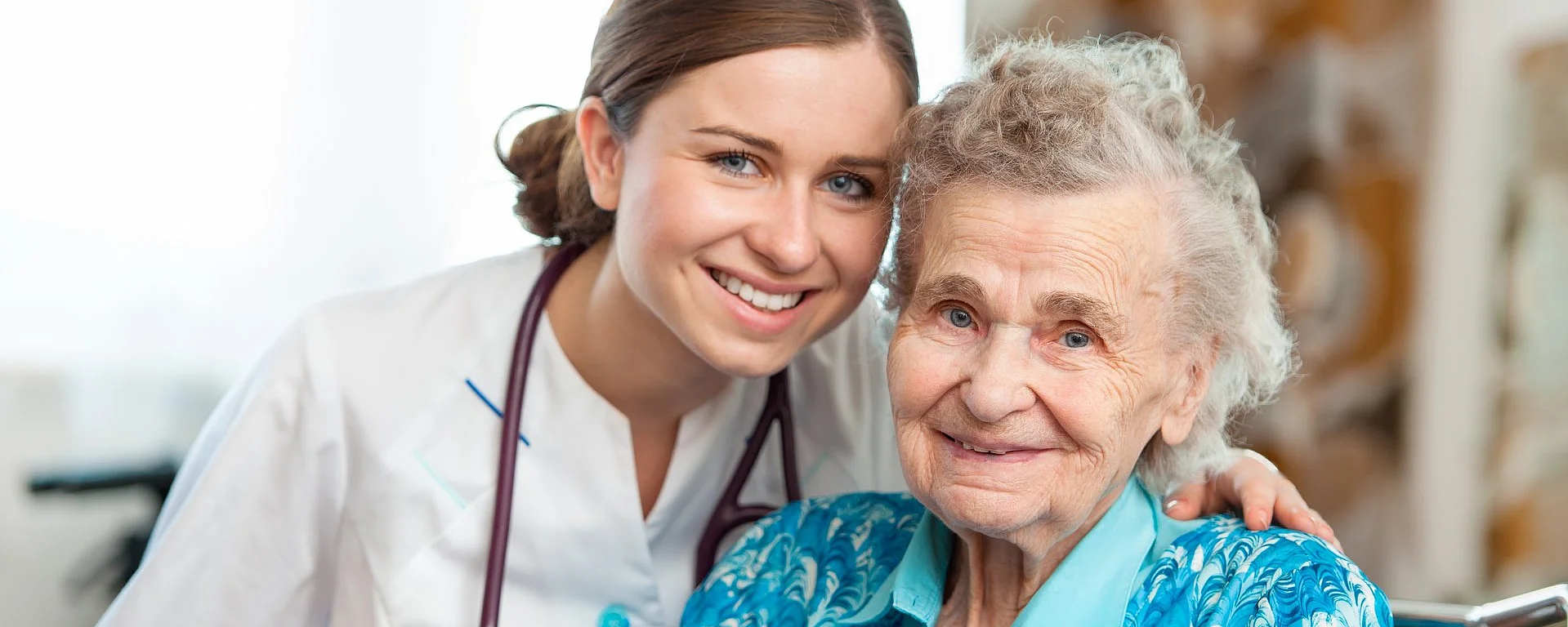 Junge Ärztin lächelt gemeinsam mit älterer Patientin im Rollstuhl in die Kamera.