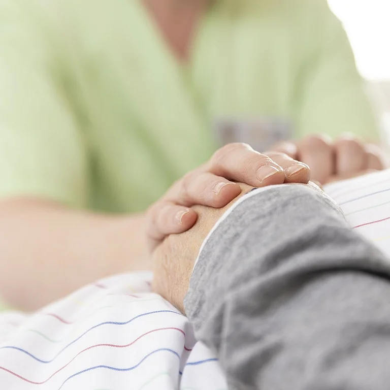 Eine Pflegekraft hält einfühlsam die Hand einer liegenden Person – ein Moment der Fürsorge und menschlichen Nähe.