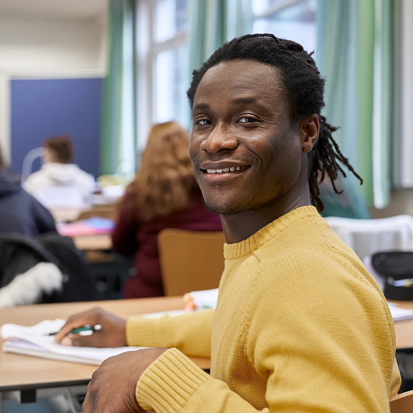Ein lächelnder Teilnehmer blickt in einem Schulungsraum zur Kamera – im Hintergrund läuft der Unterricht.