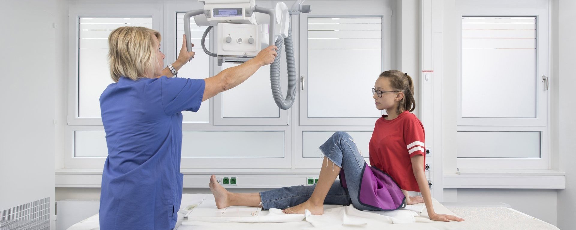 Medizinische Fachkraft positioniert ein Röntgengerät über dem Bein eines Mädchens in einem Untersuchungsraum.