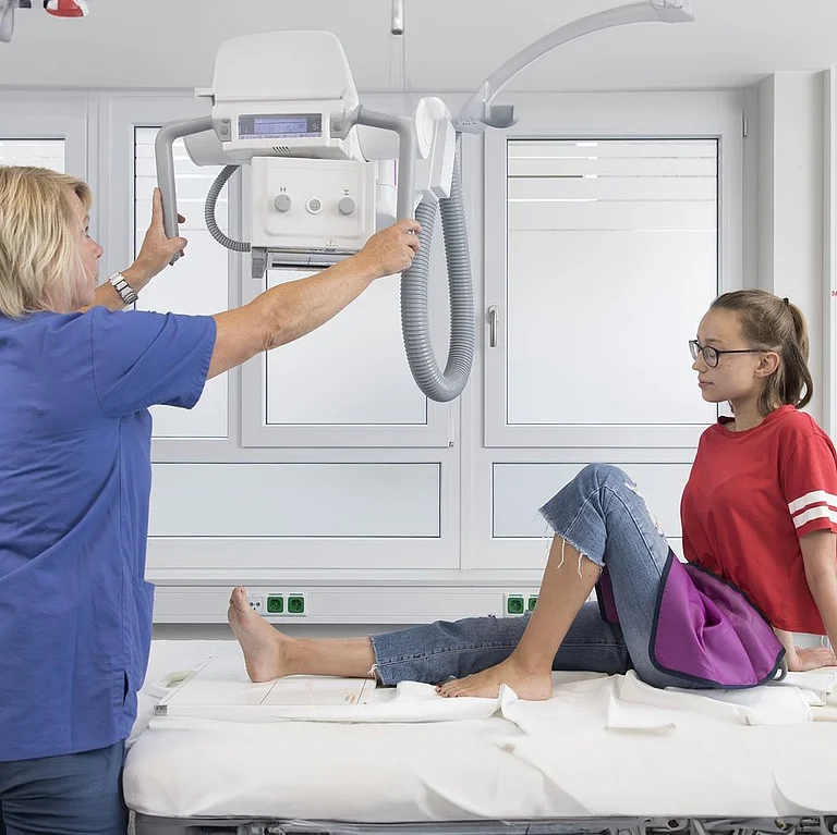 Medizinische Fachkraft positioniert ein Röntgengerät über dem Bein eines Mädchens in einem Untersuchungsraum.