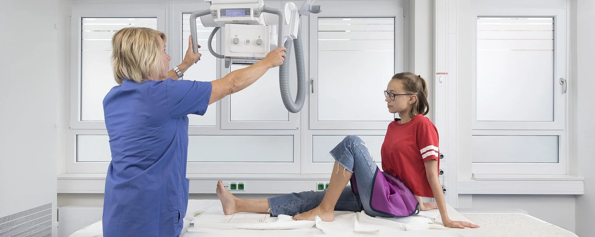 Medizinische Fachkraft positioniert ein Röntgengerät über dem Bein eines Mädchens in einem Untersuchungsraum.