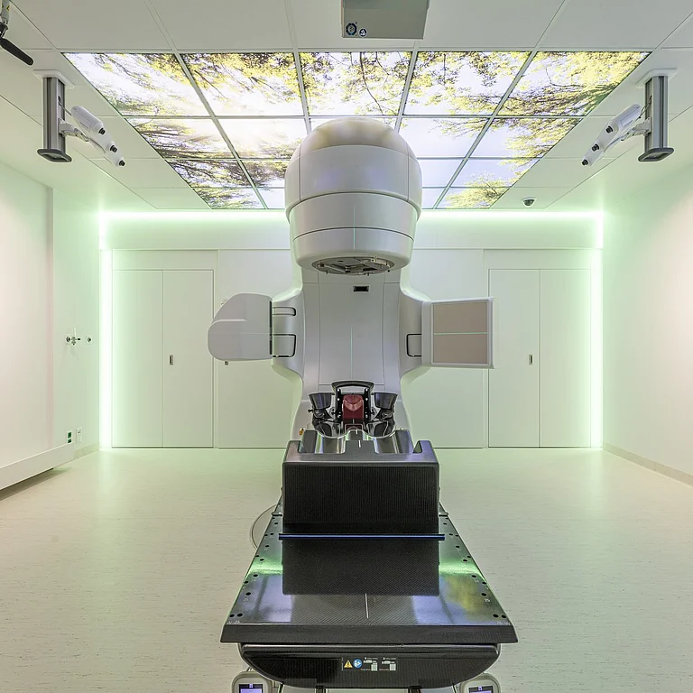 Ein Gerät zur Strahlentherapie steht in einem hellen Raum. An der Decke und den Wänden hängen Landschaftsbilder.