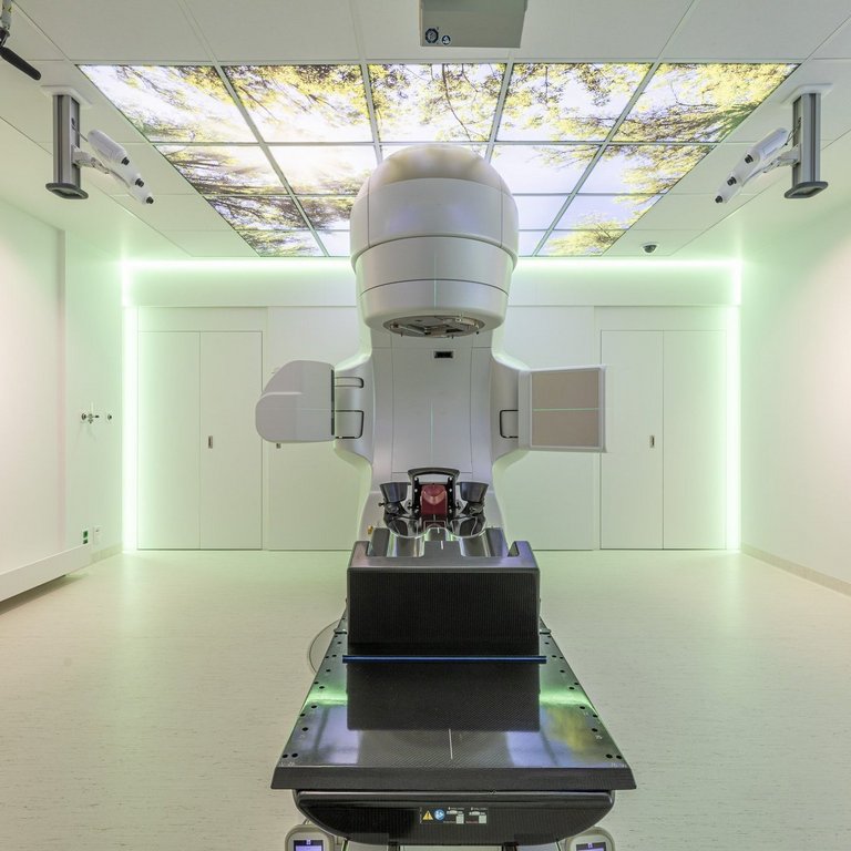Ein Gerät zur Strahlentherapie steht in einem hellen Raum. An der Decke und den Wänden hängen Landschaftsbilder.