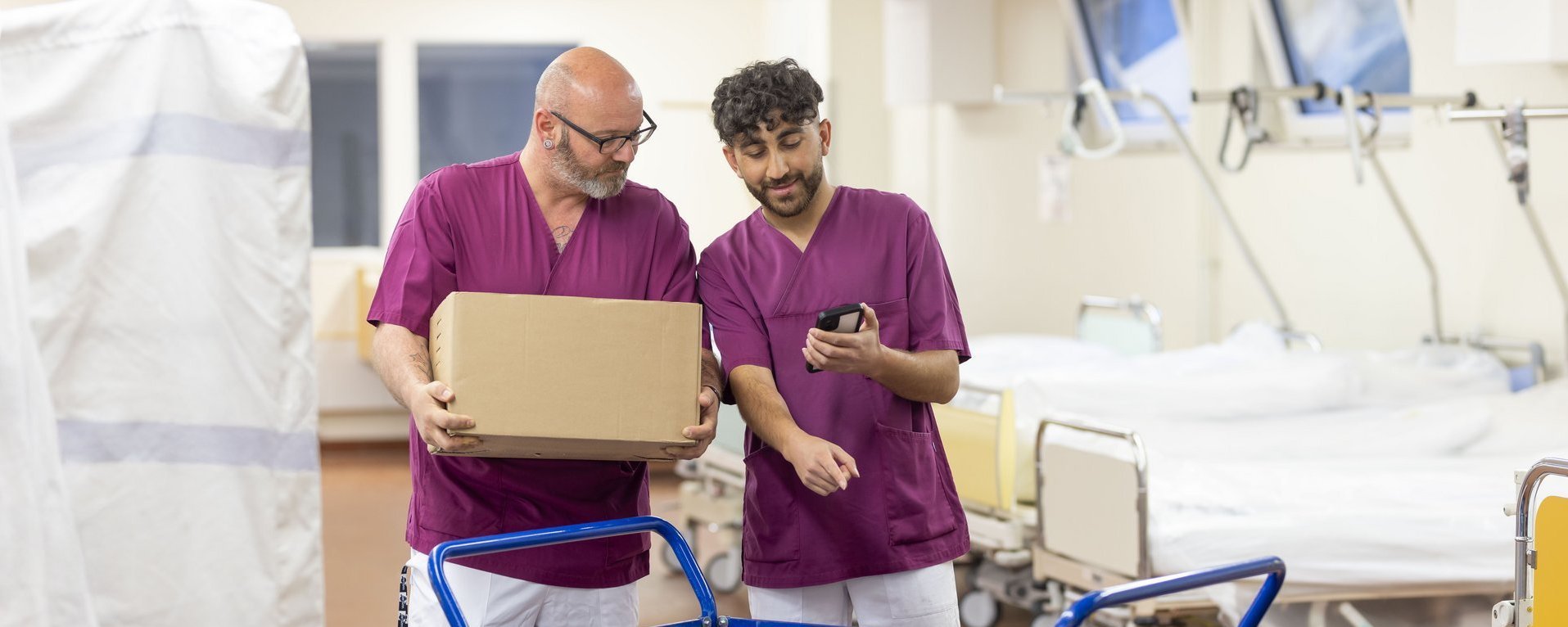 Zwei Männer in fliederfarbenen Kasacks unterhalten sich in einem Krankenhaus-Betten-Zimmer. Vor ihnen steht ein blauer Schiebewagen mit zwei Truhen. Auf dem Wagen steht das Wort „Logistik“.