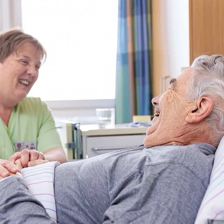 Pflegerin hält die Hand eines älteren Patienten im Krankenbett und lächelt ihm zu.