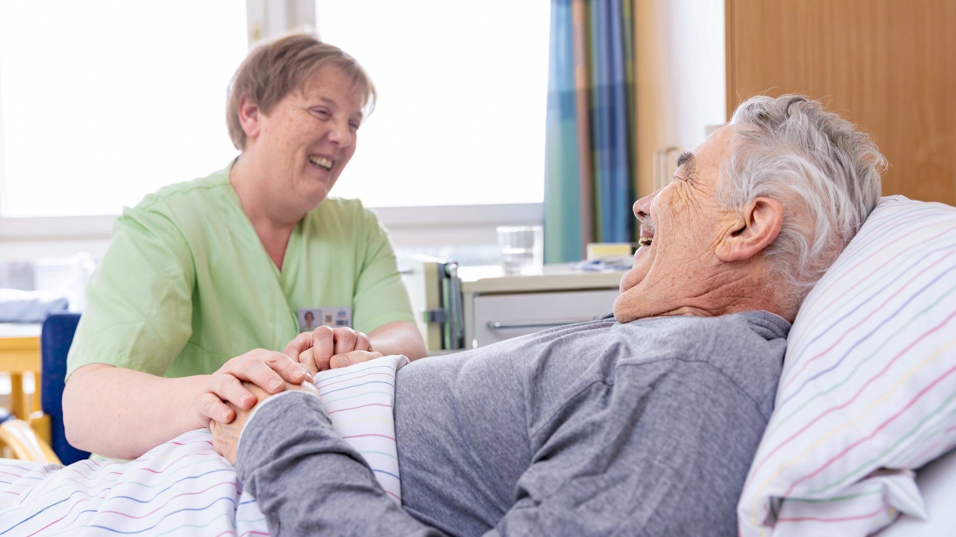 Pflegerin hält die Hand eines älteren Patienten im Krankenbett und lächelt ihm zu.