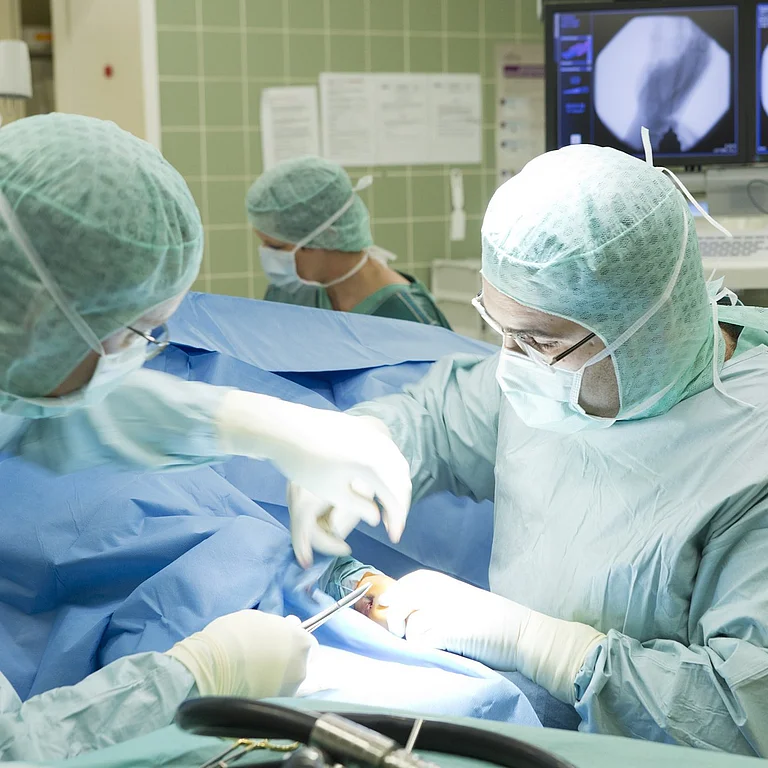 Zwei Chirurg*innen operieren im OP-Saal, vollständig in OP-Kleidung. Im Hintergrund sind Monitore mit Röntgenbildern zu sehen.