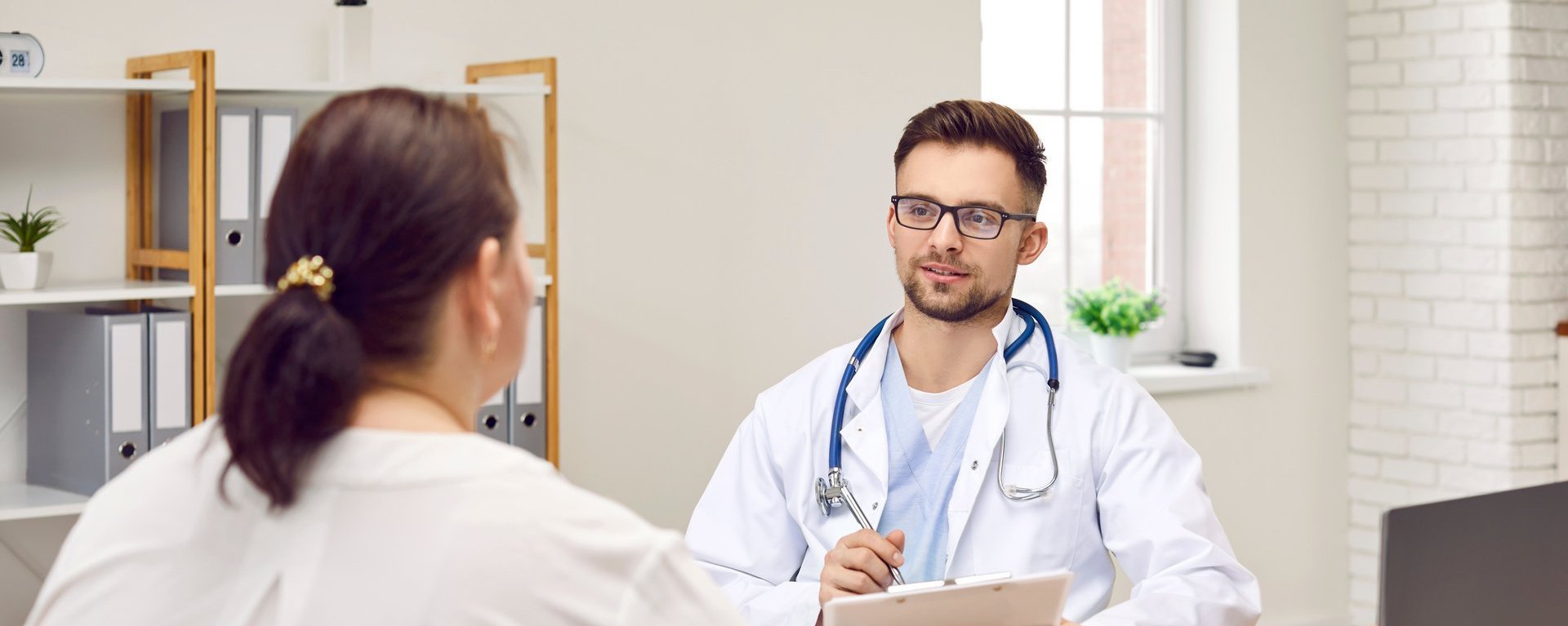 Ein Arzt mit Stethoskop und Klemmbrett spricht mit einer Patientin in einem Büro.