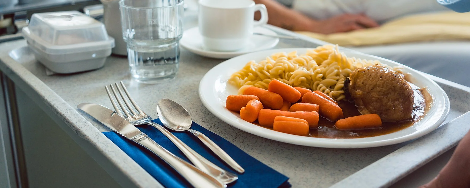 Essen wird einem Patienten im Krankenhaus ans Bett serviert. Der Fokus liegt auf dem Essen, das aus Karotten, Nudeln, Fleisch und dunkler Sauce besteht.