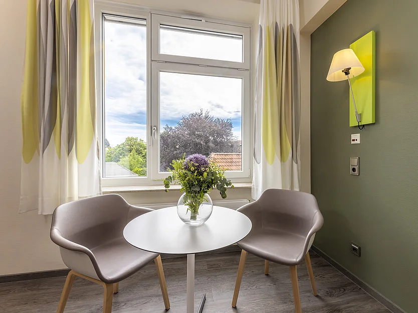 Sitzbereich mit zwei Stühlen und rundem Tisch vor einem Fenster mit Ausblick ins Grüne.