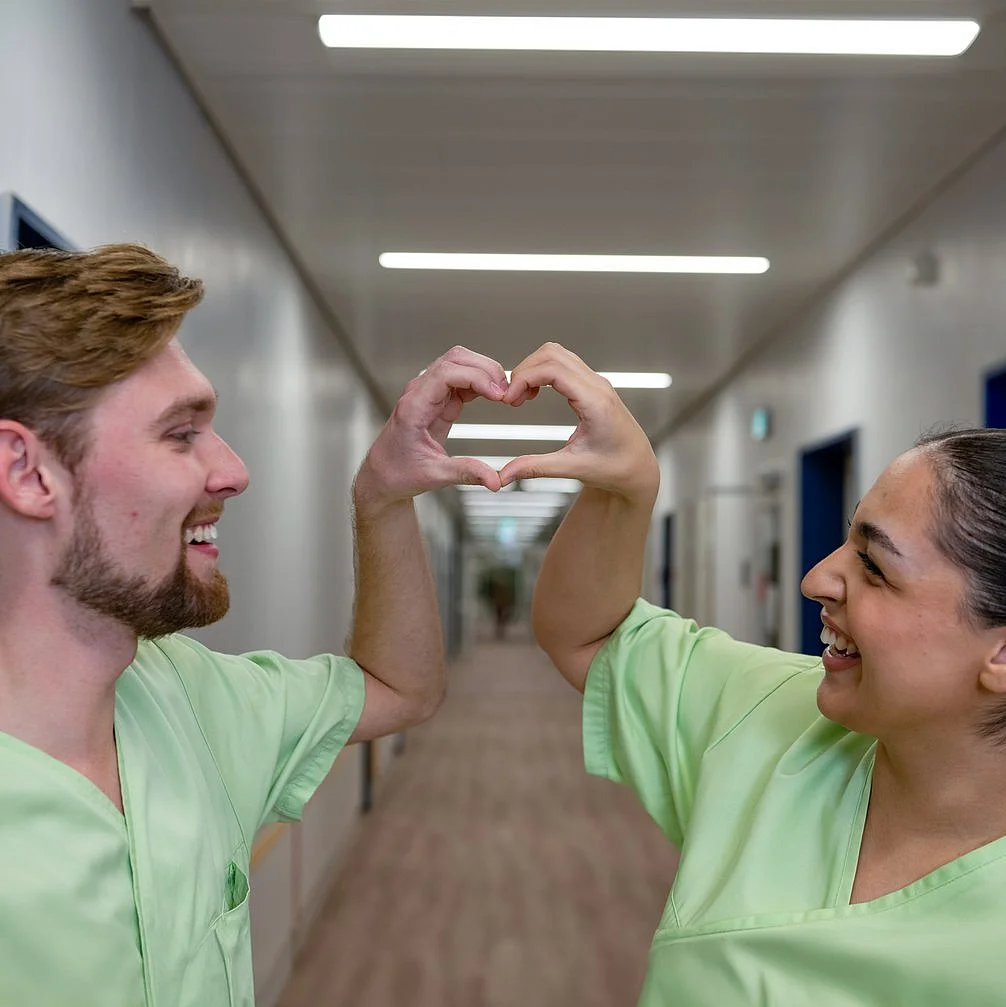 Zwei junge Pflegekräfte formen mit ihren Händen ein Herzsymbol in einem Krankenhausflur.