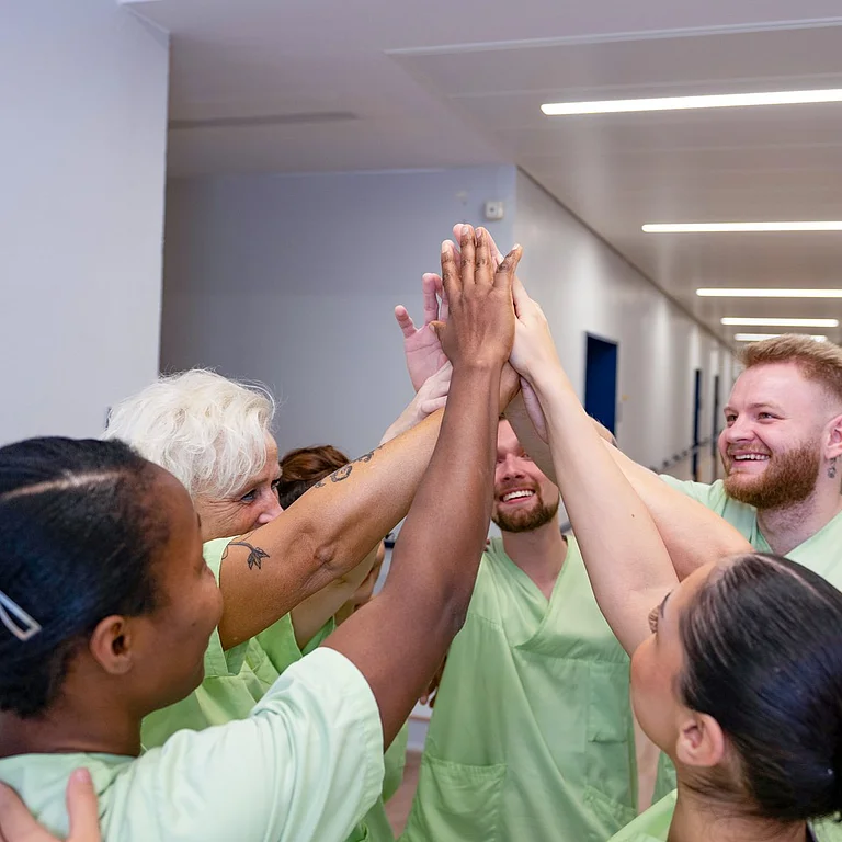 Pflegeteam steht im Kreis und legt im Flur motiviert die Hände zu einem High Five zusammen.