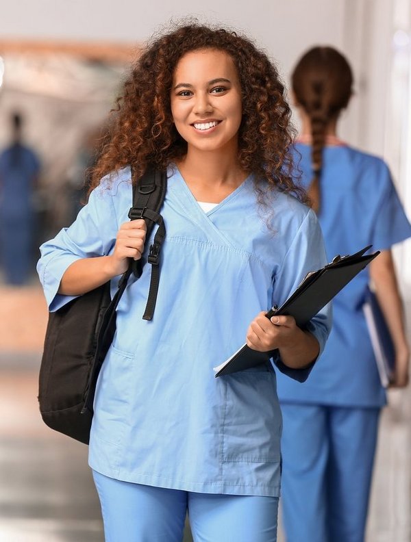 Junge Medizinstudentin mit Rucksack und Unterlagen lächelt in die Kamera – im Hintergrund weitere Studierende in blauen Kitteln auf dem Klinikflur.