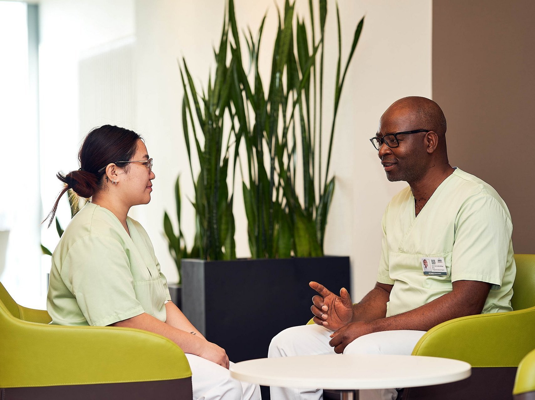 Zwei Pflegekräfte in grüner Kleidung sitzen sich in einer modernen, hellen Sitzecke gegenüber und unterhalten sich freundlich. Im Hintergrund steht eine große Zimmerpflanze.