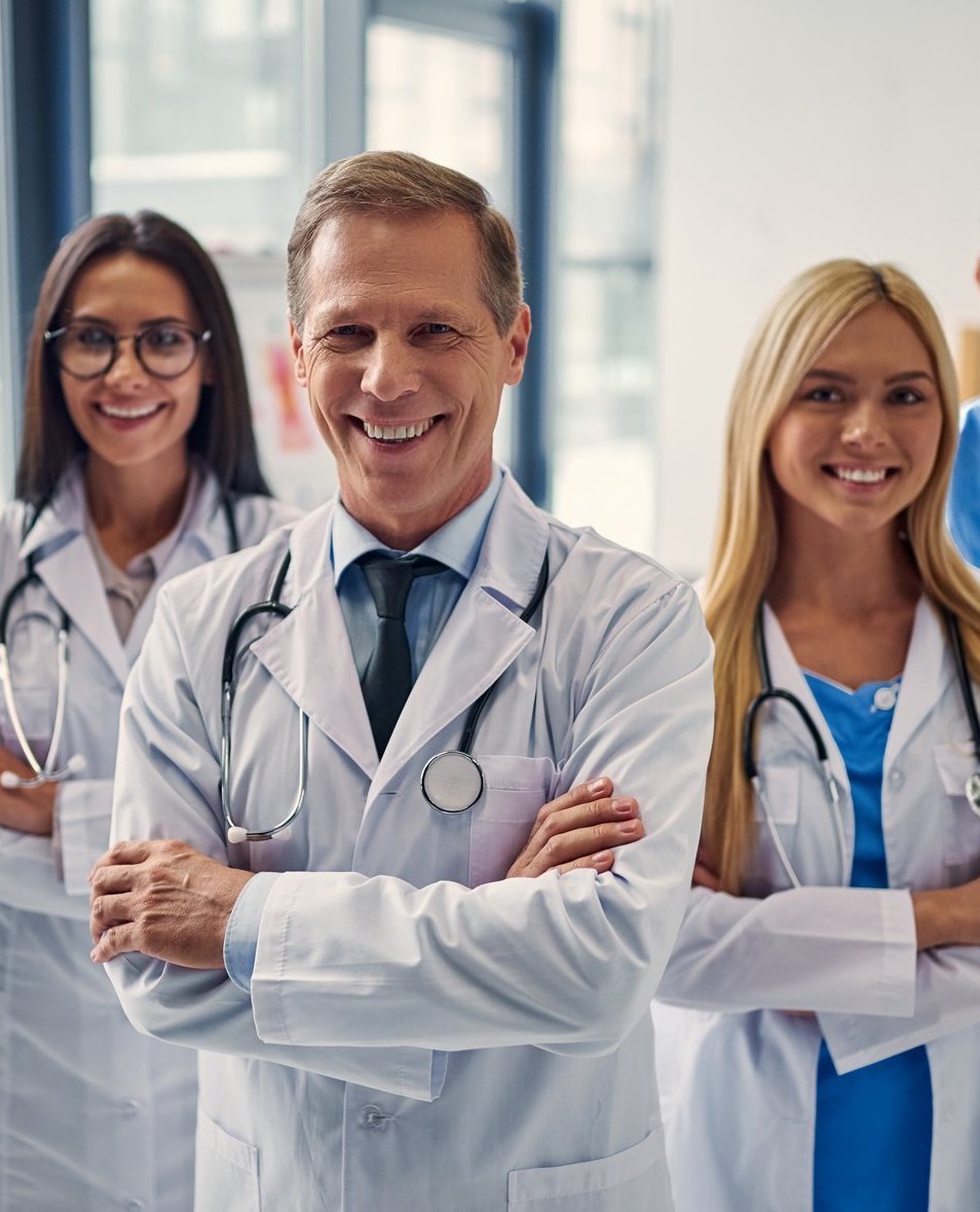 Lächelndes medizinisches Team mit Ärzten und Ärztinnen in weißem Kittel und Pflegekraft in blauer Kleidung im Klinikflur.