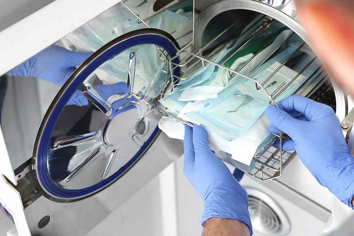 Eine Person mit blauen Handschuhen entnimmt steril verpackte medizinische Instrumente aus einem Autoklaven. Die Szene zeigt einen Arbeitsschritt im Bereich der Instrumentenaufbereitung zur Sicherstellung der Hygiene.