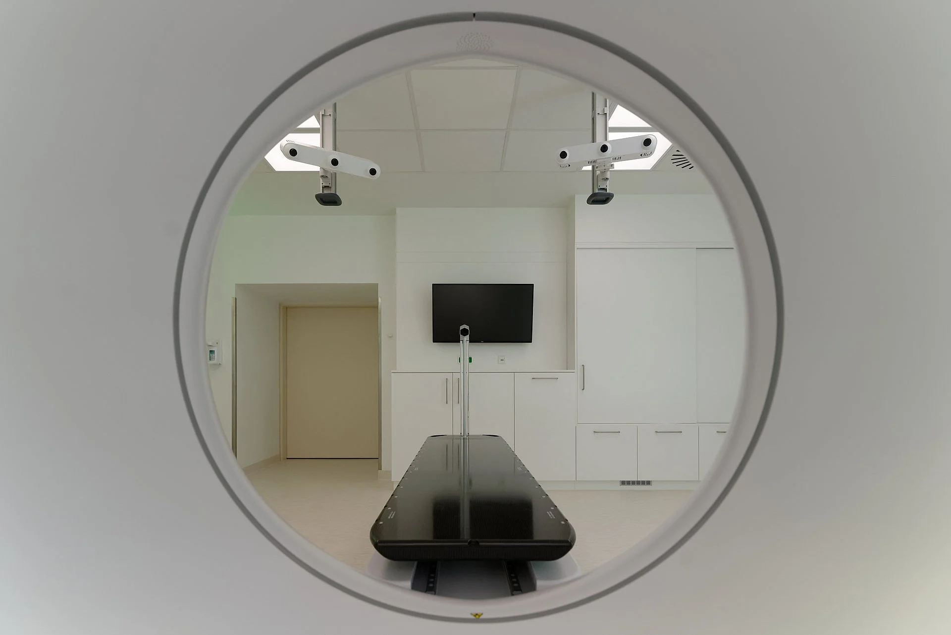 Blick durch die Öffnung eines Strahlentherapiegeräts auf einen leeren, hellen Behandlungsraum mit Monitor.