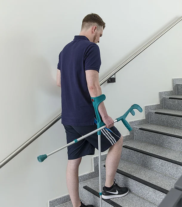 Junger Mann steigt mit Unterarmgehstützen eine Treppe hinauf und hält sich am Geländer fest.