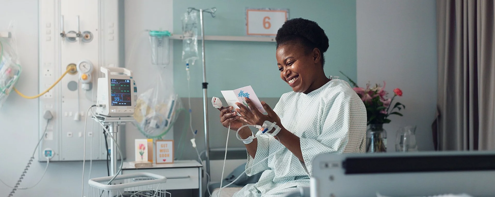 Eine Frau im Krankenhauskittel sitzt in einem Bett im Krankenhaus und sieht sich eine Grußkarte an, die sie in der Hand hält.