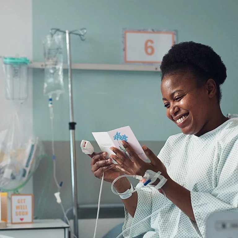 Eine Frau im Krankenhauskittel sitzt in einem Bett im Krankenhaus und sieht sich eine Grußkarte an, die sie in der Hand hält.