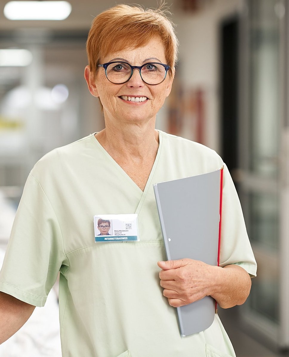 Pflegekraft mit Brille steht lächelnd auf einer Krankenhausstation und hält eine Patientenakte in der Hand.