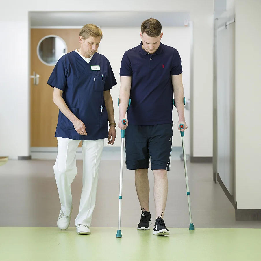 Therapeut begleitet einen jungen Mann mit Unterarmgehstützen beim Gehen im Klinikflur.