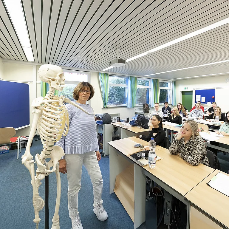 Die Szene zeigt einen lebendigen Unterricht im medizinischen Bereich: Eine Lehrkraft erklärt anatomische Zusammenhänge am Skelettmodell, während die Studierenden aufmerksam folgen.