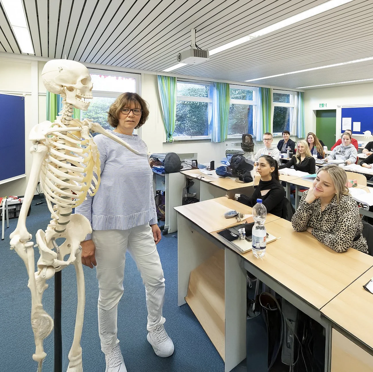 Die Szene zeigt einen lebendigen Unterricht im medizinischen Bereich: Eine Lehrkraft erklärt anatomische Zusammenhänge am Skelettmodell, während die Studierenden aufmerksam folgen.