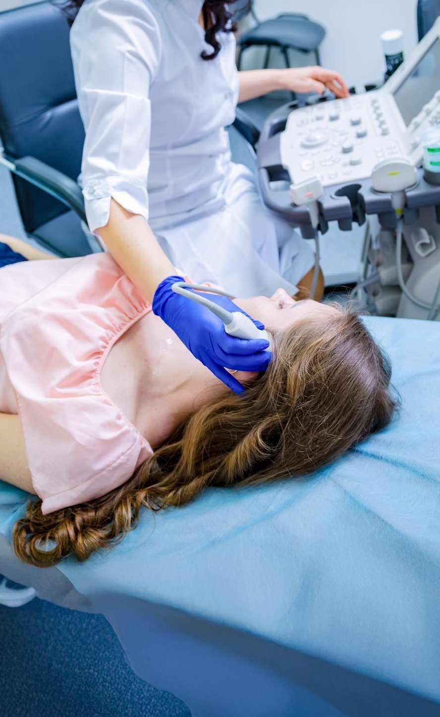 Eine Patientin liegt auf einer Untersuchungsliege, während eine Ärztin mit einem Ultraschallgerät eine Untersuchung im Halsbereich durchführt.