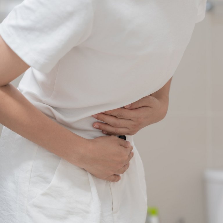 Eine Frau in weißer Kleidung hält sich ihren schmerzenden Bauch. Im Hintergrund ist eine Toilette zu sehen.