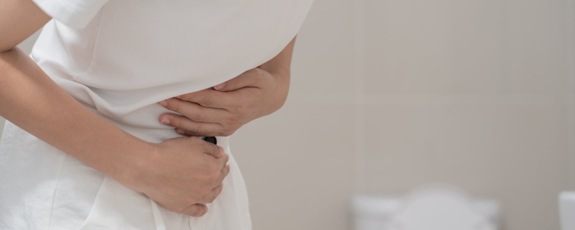 Eine Frau in weißer Kleidung hält sich ihren schmerzenden Bauch. Im Hintergrund ist eine Toilette zu sehen.