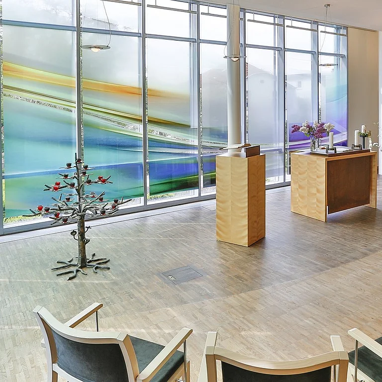 Blick von hinten in die Kapelle des Allgemeinen Krankenhaus Celle. Man sieht getönte, bunte Scheiben, einen Altar, Ambo und Stühle für Besucher.