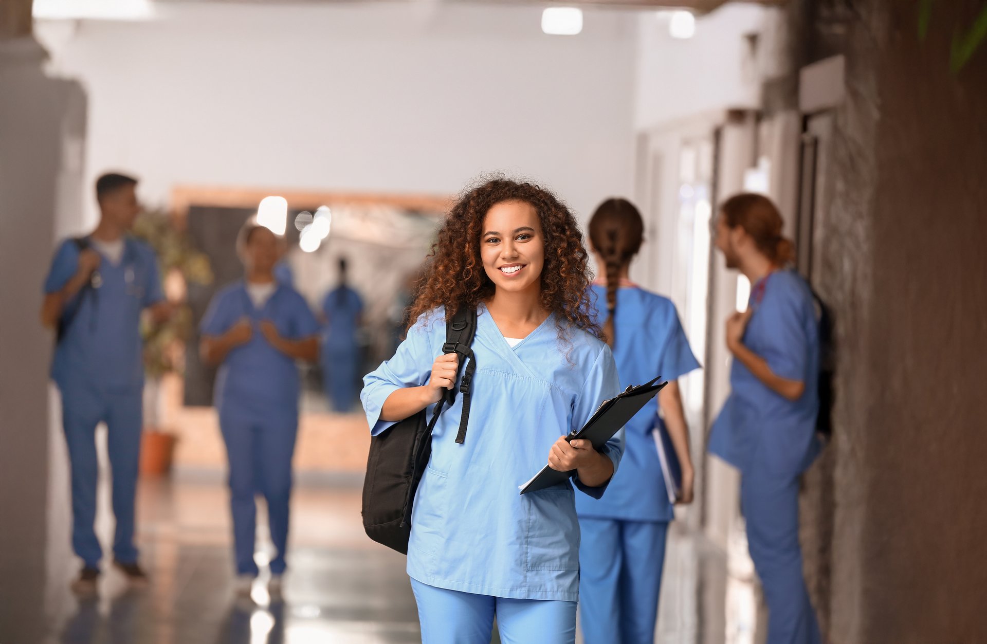 Junge Medizinstudentin mit Rucksack und Unterlagen lächelt in die Kamera – im Hintergrund weitere Studierende in blauen Kitteln auf dem Klinikflur.